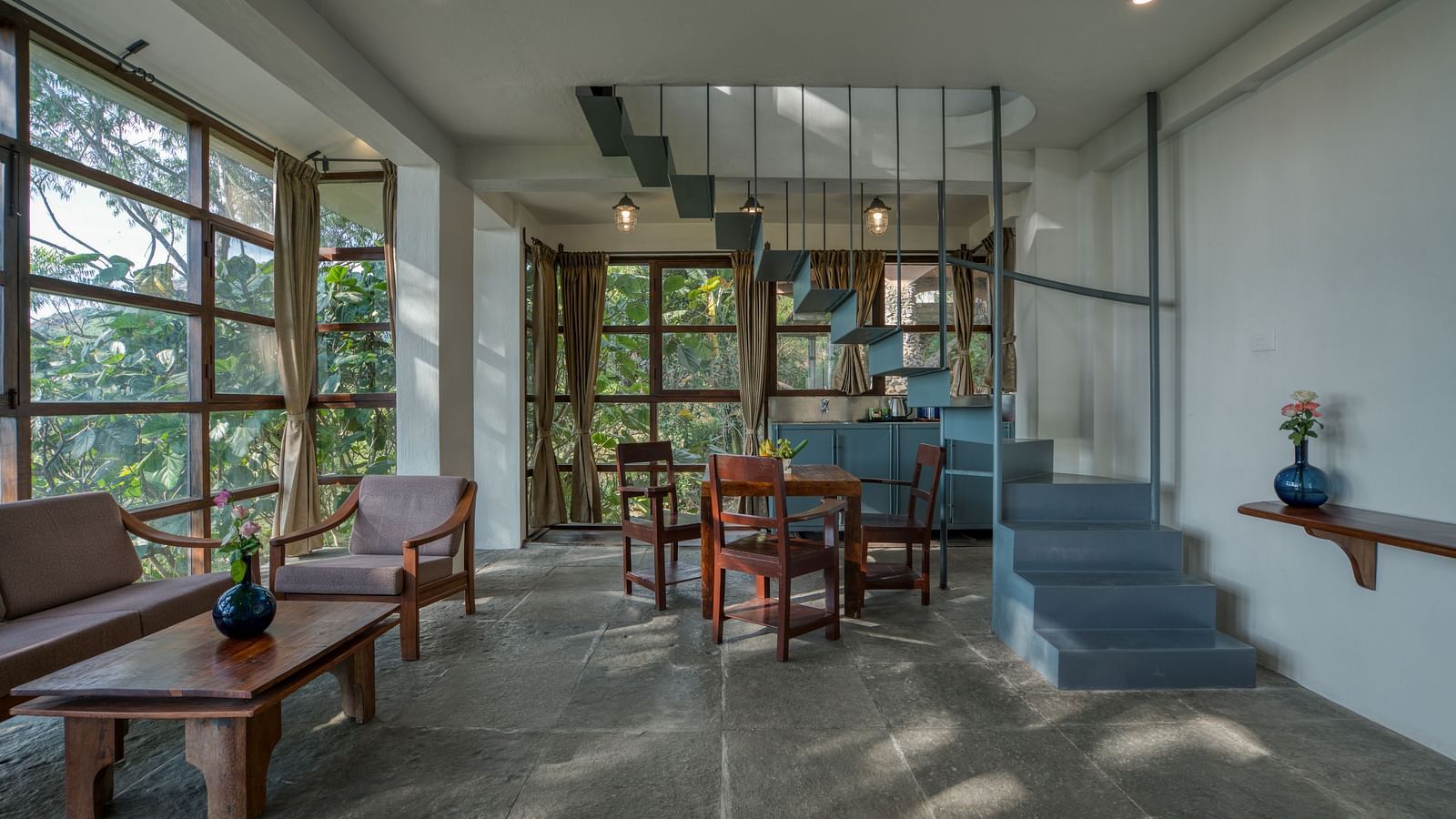 Spacious living area with a blue staircase, wooden furniture, and expansive windows providing a view of the lush greenery outside. | Mango Hill, Yercaud