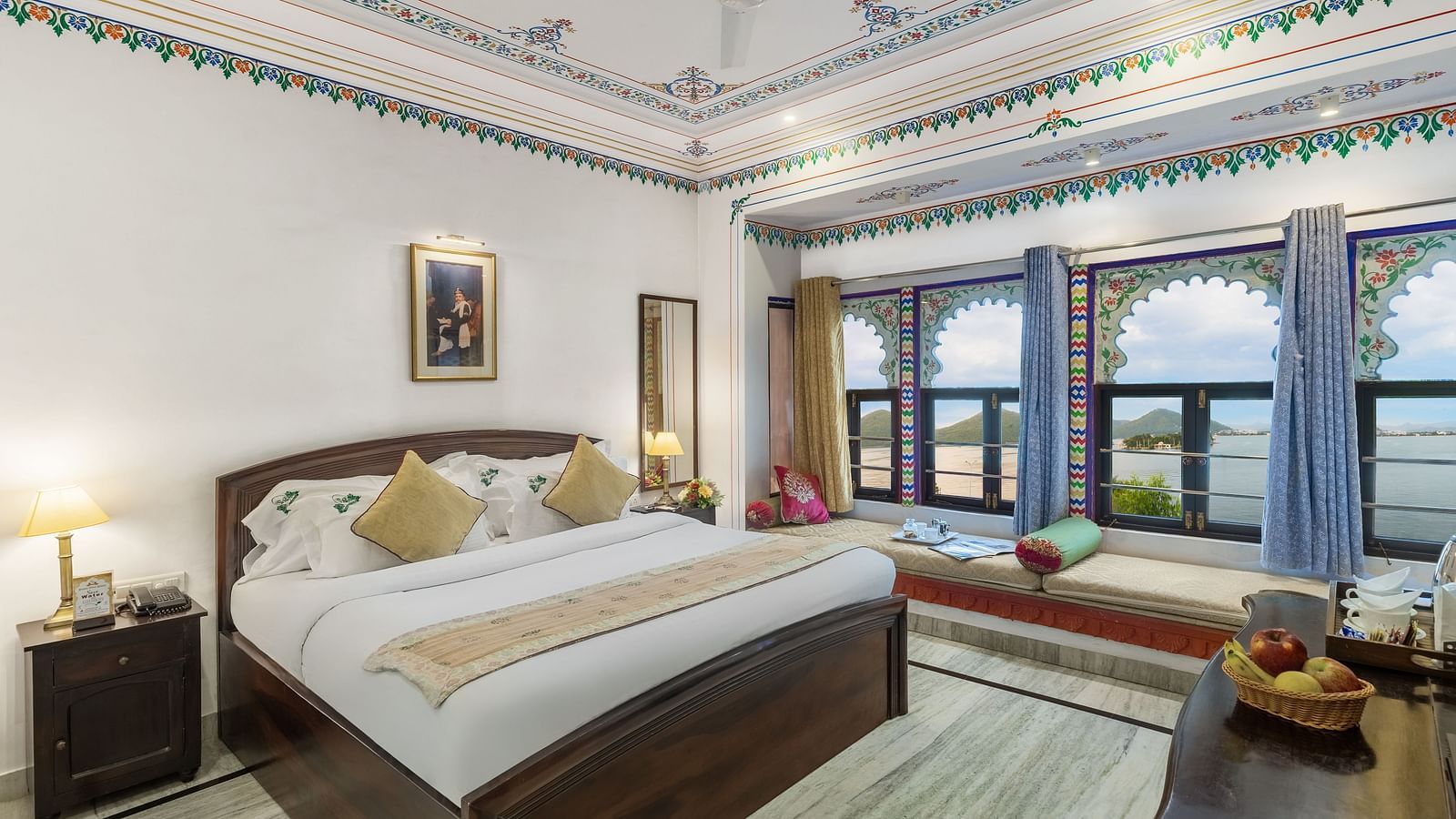  A spacious bed with a wooden bedside table and a label, alongside bay window seating with curtains pulled to the side at Panna Vilas Palace