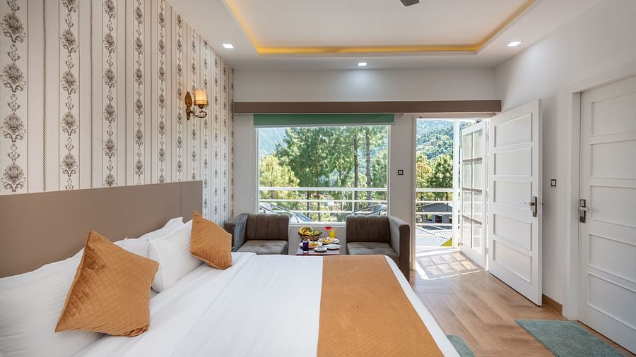 Bright hotel room with a comfortable double bed dressed in white and ochre linen, patterned wallpaper, a sitting area by the window, and French doors opening to a balcony with serene views of trees and distant hills - DLS Nature Trinket Resort, Dalhousie.