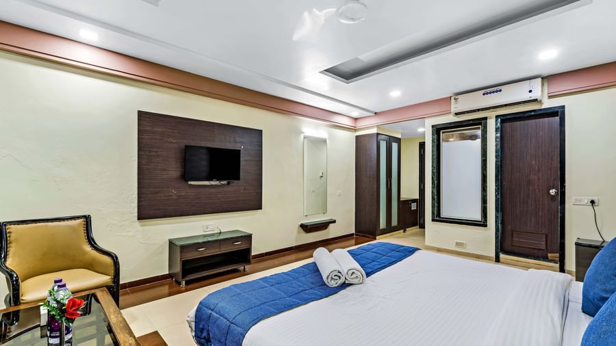 A wide view of a suite in Krushnai Resort featuring a double bed with blue runner, a wall mounted TV, modern furnishings and a lounge chair.