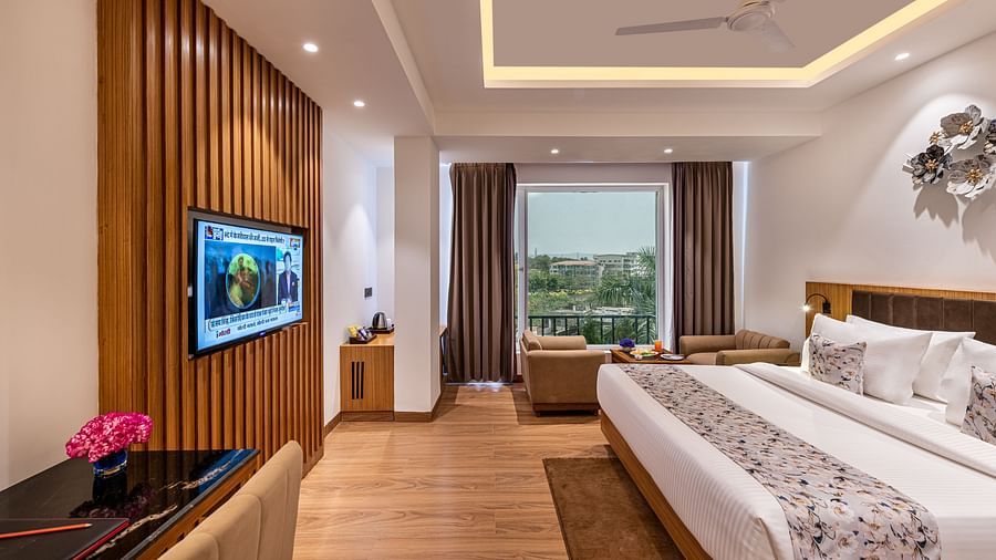 A hotel room with a king size bed, a flat-screen TV on a wood-panelled wall, and a city view through expansive windows. It features a distinctive tray ceiling with lighting, a ceiling fan, and floor-to-ceiling curtains - Lords Eco Inn Dehradun.