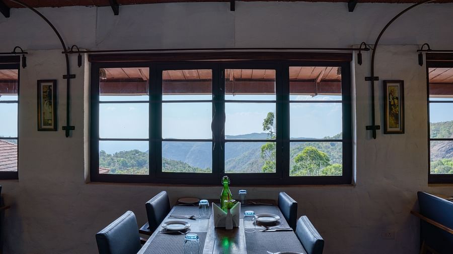 Dining table set near large windows with a scenic view of the hills and greenery. | Mango Hill, Yercaud