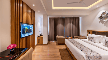 An elegant designed spacious room with a king size bed, wall-mounted TV, work desk with a pink flower, seating area near full-length curtains, and wooden flooring at Lords Eco Inn Dehradun.