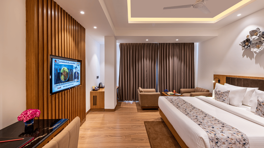 An elegant designed spacious room with a king size bed, wall-mounted TV, work desk with a pink flower, seating area near full-length curtains, and wooden flooring at Lords Eco Inn Dehradun.