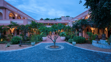 A beautiful hotel courtyard at dusk, with a paved driveway, lush green plants, and pink-colored buildings with arched walkways.