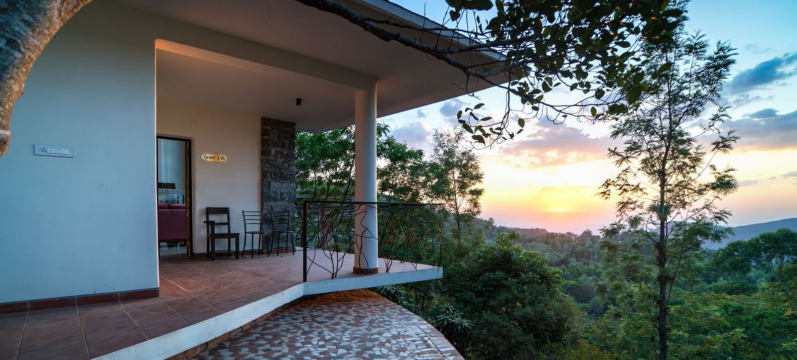 A balcony offering a stunning view of the sunset over a lush green landscape, with a table and chairs for relaxation.  | Mango Hill, Yercaud