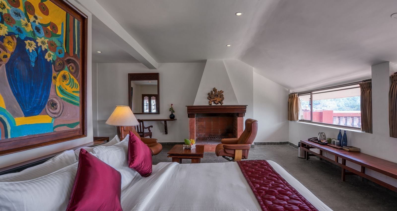 Spacious hotel room with a cozy bed, colorful wall art, fireplace, and large windows. | Mango Hill, Yercaud