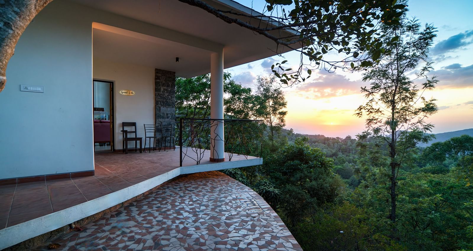 A balcony offering a stunning view of the sunset over a lush green landscape, with a table and chairs for relaxation.  | Mango Hill, Yercaud