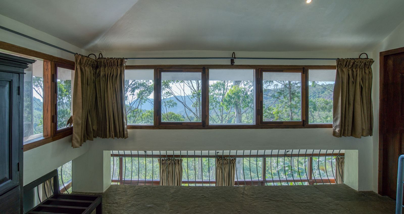 Room with large windows offering a panoramic view, furnished with simple wooden furniture. | Mango Hill, Yercaud