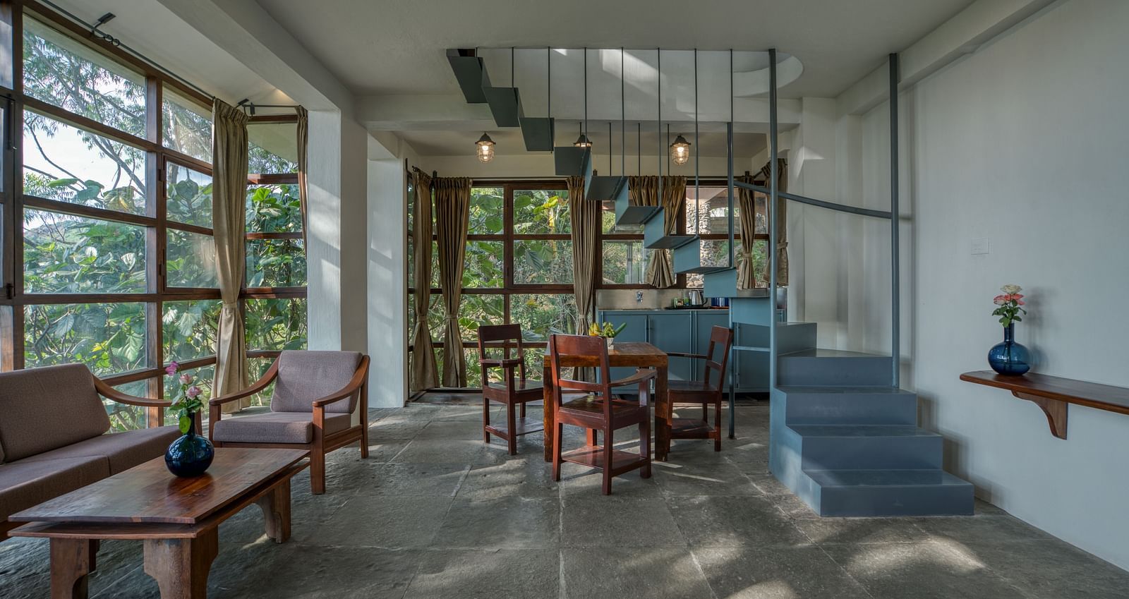 Spacious living area with a blue staircase, wooden furniture, and expansive windows providing a view of the lush greenery outside. | Mango Hill, Yercaud