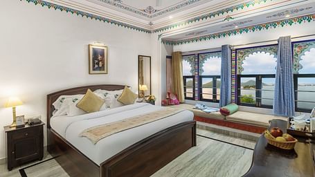  A spacious bed with a wooden bedside table and a label, alongside bay window seating with curtains pulled to the side at Panna Vilas Palace