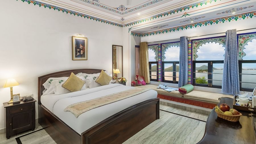  A spacious bed with a wooden bedside table and a label, alongside bay window seating with curtains pulled to the side at Panna Vilas Palace
