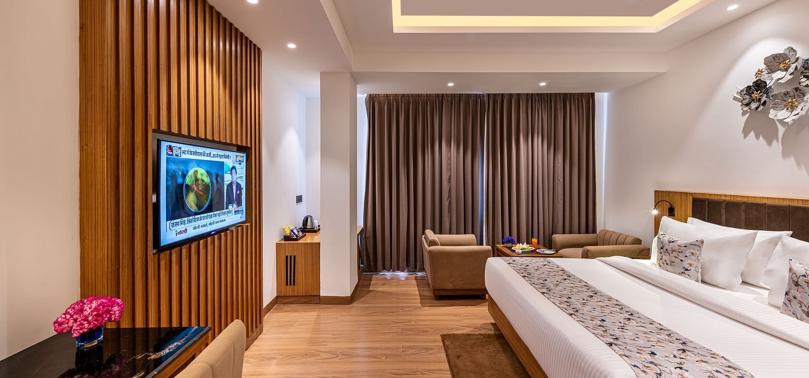 Spacious hotel room with a king size bed, wall-mounted TV, work desk with a pink flower, seating area near full-length curtains, and warm wooden flooring with modern lighting - Lords Eco Inn Dehradun.