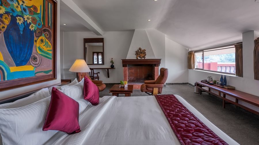Spacious hotel room with a cozy bed, colorful wall art, fireplace, and large windows. | Mango Hill, Yercaud