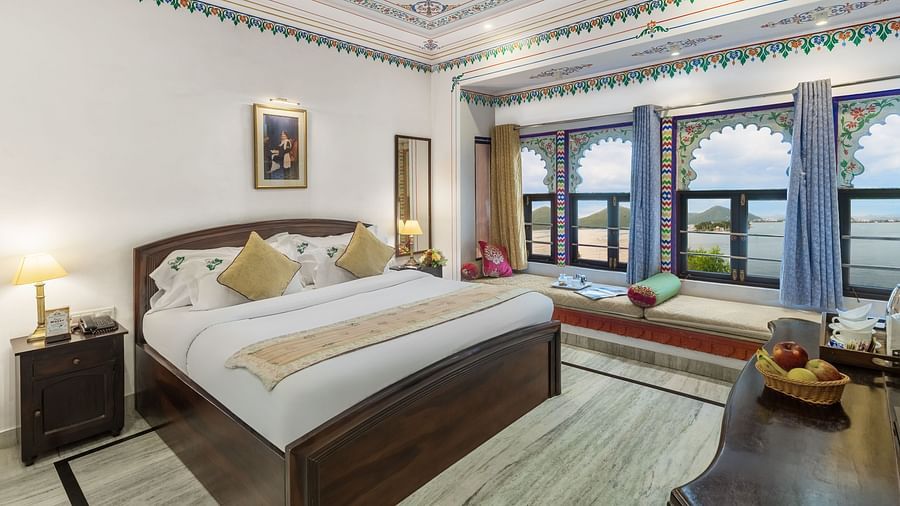  A spacious bed with a wooden bedside table and a label, alongside bay window seating with curtains pulled to the side at Panna Vilas Palace