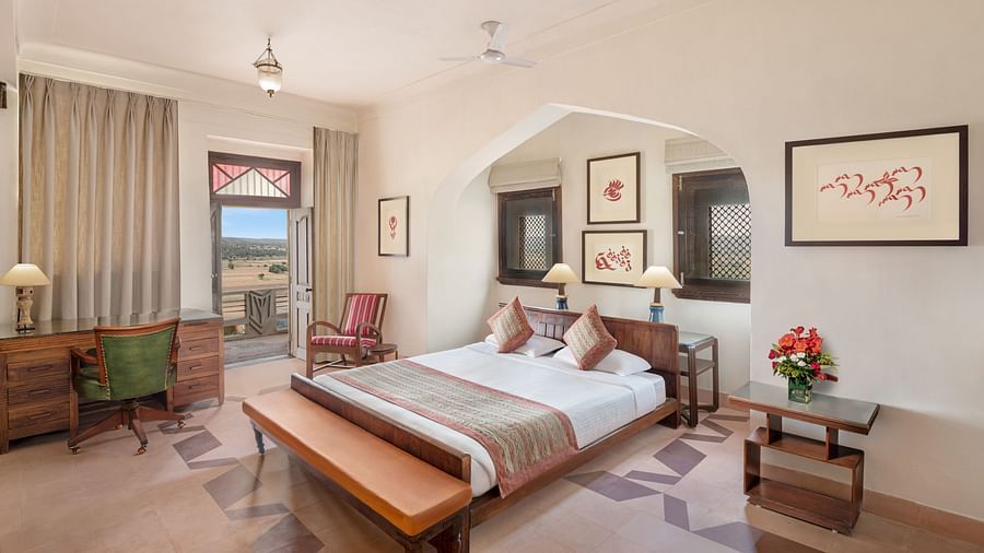 A room with a bed, a desk, a chair, a bench, a small table with flowers, and framed artworks, with a doorway leading to a balcony in Tijara Fort-Palace - 19th Century, Alwar.