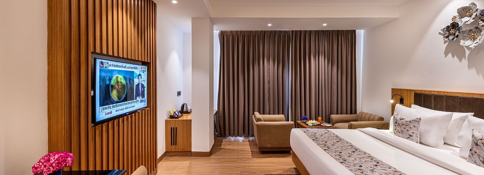 Spacious hotel room with a king size bed, wall-mounted TV, work desk with a pink flower, seating area near full-length curtains, and warm wooden flooring with modern lighting - Lords Eco Inn Dehradun.