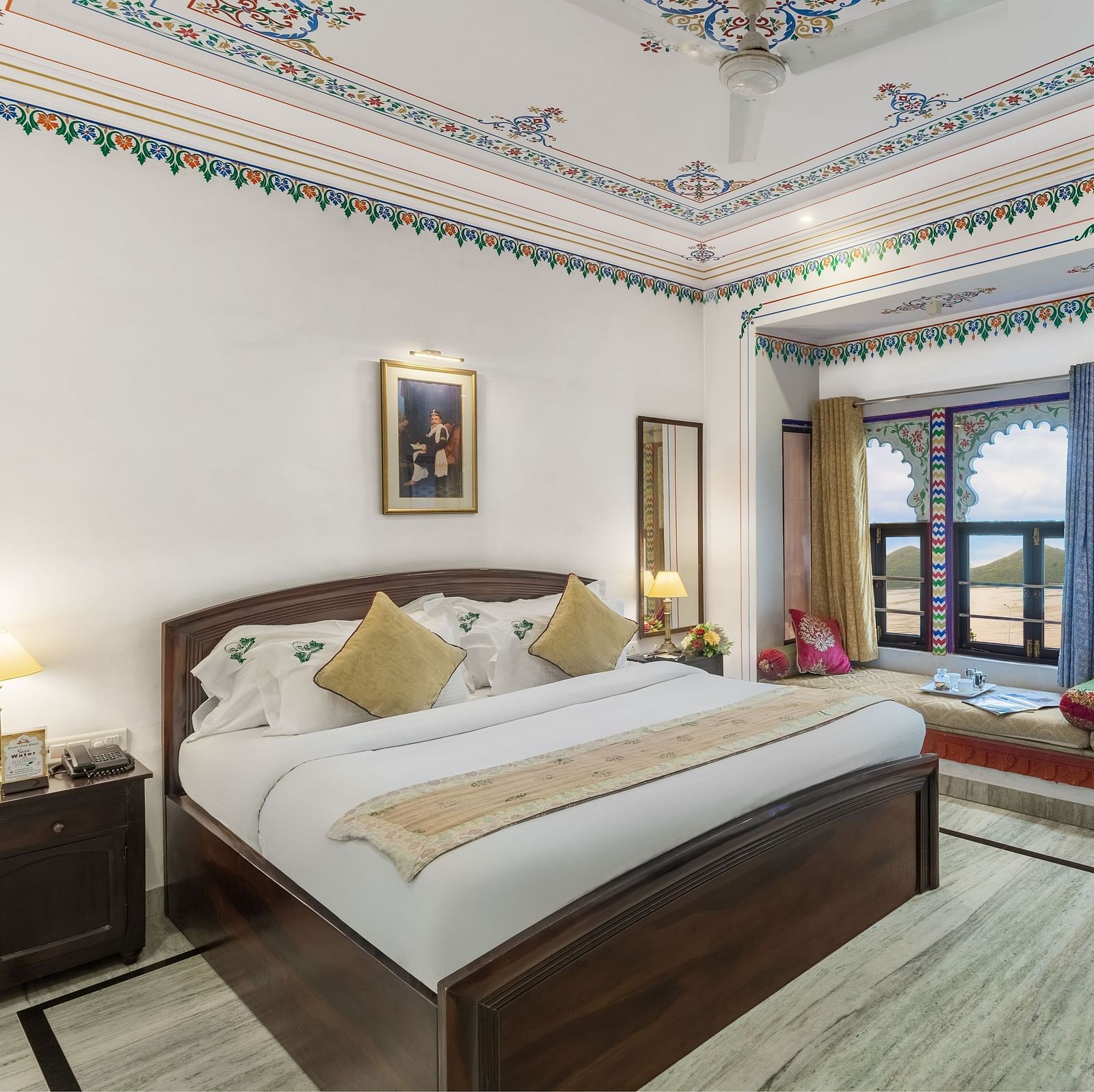  A spacious bed with a wooden bedside table and a label, alongside bay window seating with curtains pulled to the side at Panna Vilas Palace