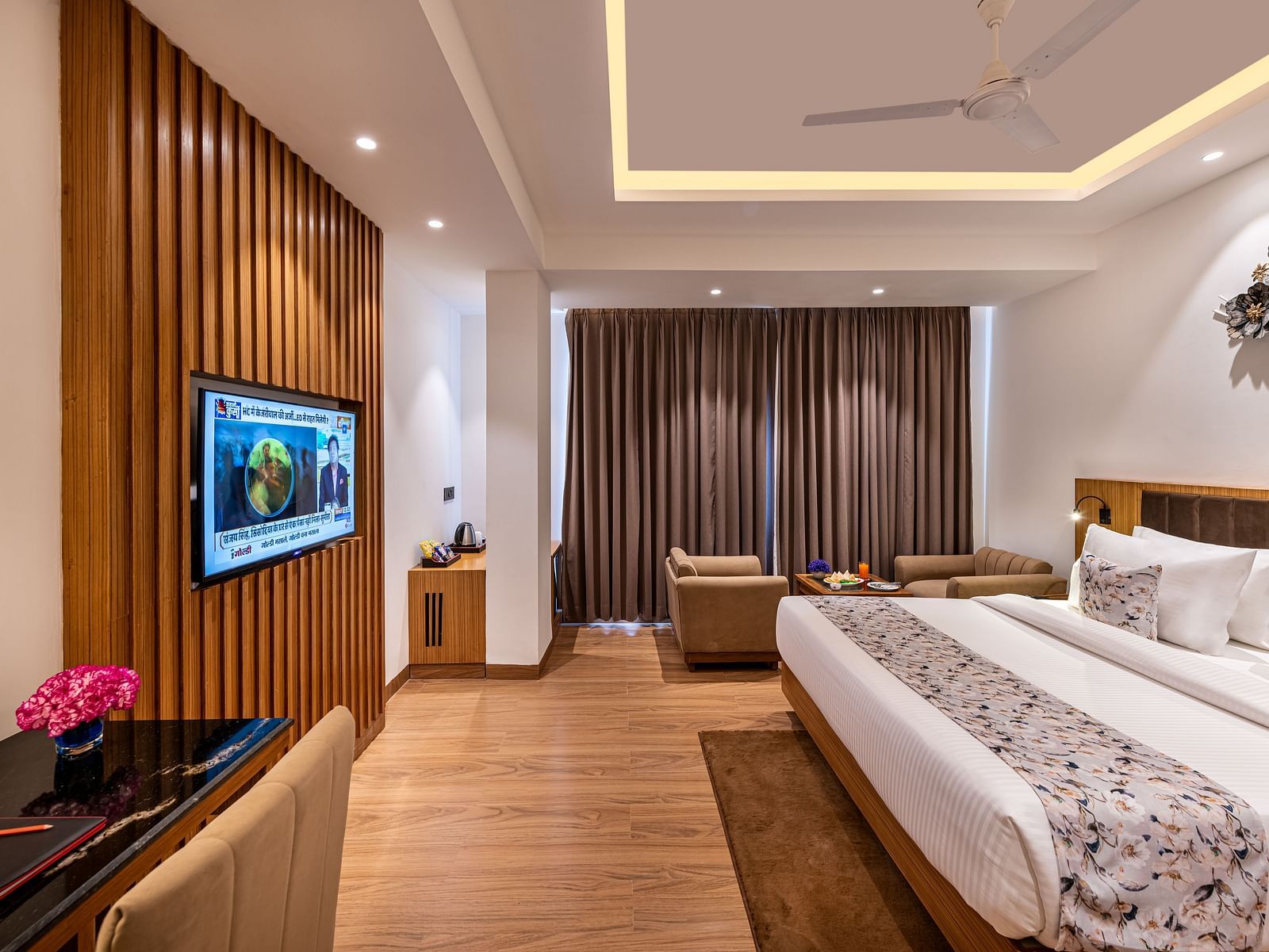 Spacious hotel room with a king size bed, wall-mounted TV, work desk with a pink flower, seating area near full-length curtains, and warm wooden flooring with modern lighting - Lords Eco Inn Dehradun.