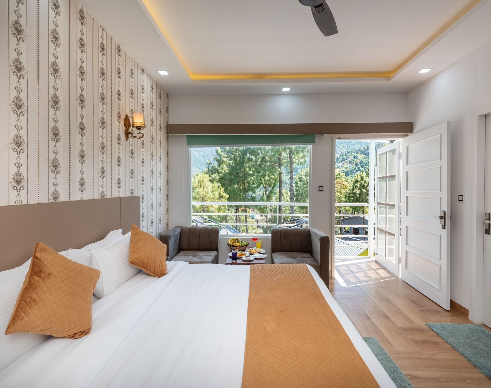 Bright hotel room with a comfortable double bed dressed in white and ochre linen, patterned wallpaper, a sitting area by the window, and French doors opening to a balcony with serene views of trees and distant hills - DLS Nature Trinket Resort, Dalhousie.