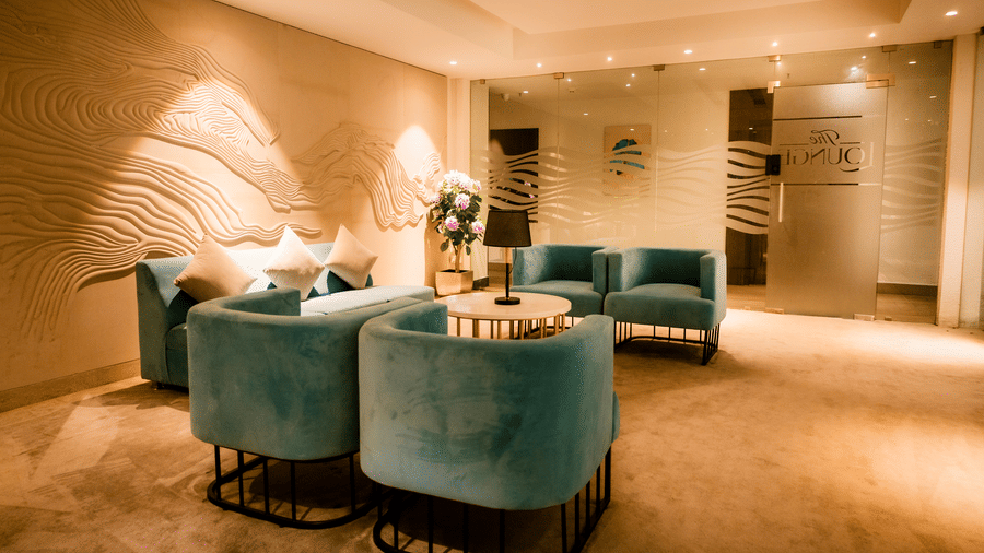 Business lounge seating with teal velvet sofas and tub chairs, a round gold-based table and a sculpted wave-panel wall at Ilara Hotels & Spa, Tada.