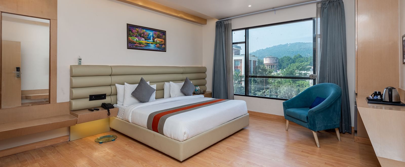A room with a bed, bed side tables, a chair, and an expansive glass window overlooking the city view - DLS Hotel  Arihant, Haridwar