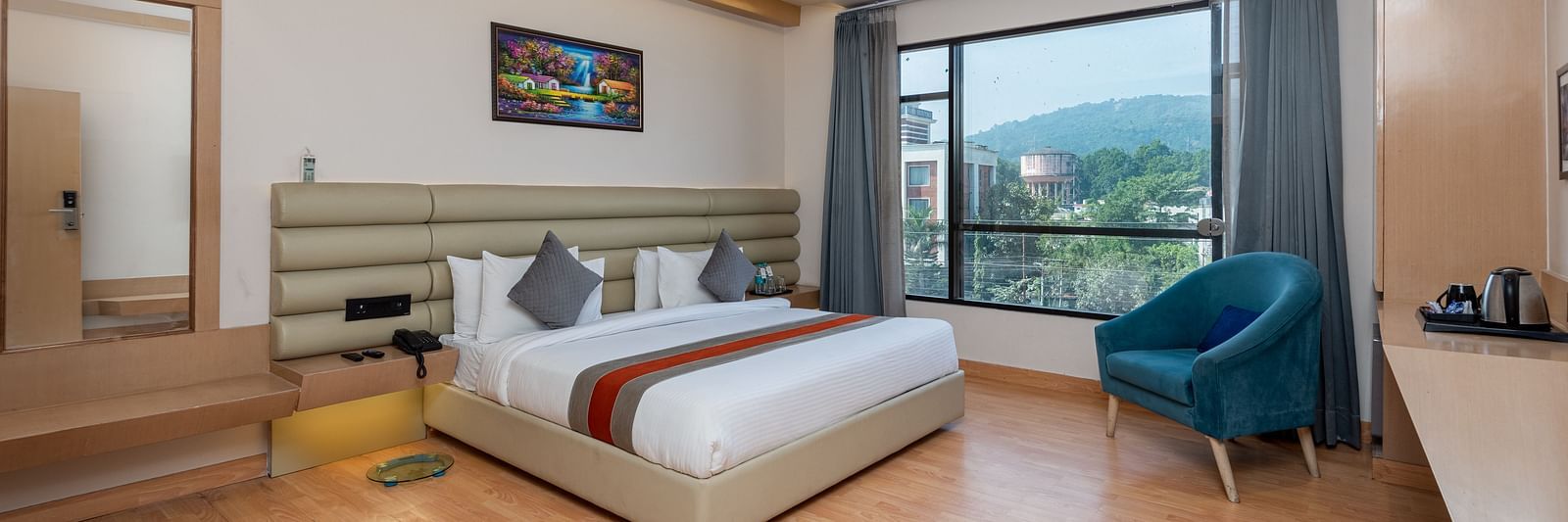 A room with a bed, bed side tables, a chair, and an expansive glass window overlooking the city view - DLS Hotel  Arihant, Haridwar