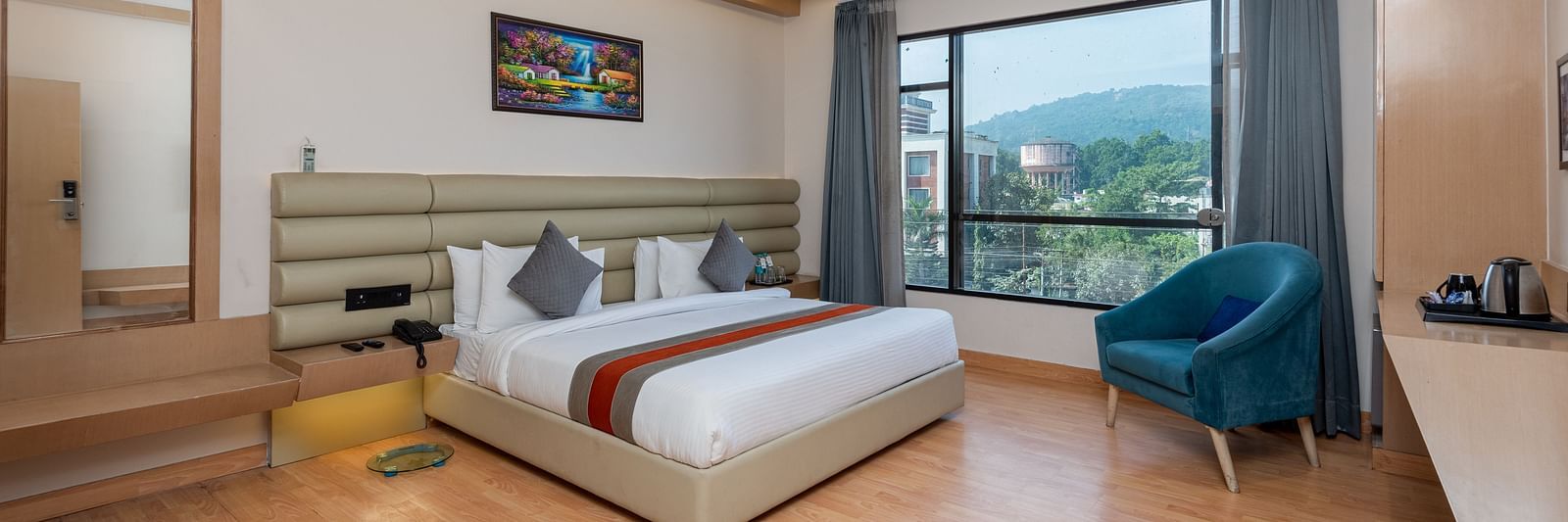 A room with a bed, bed side tables, a chair, and an expansive glass window overlooking the city view - DLS Hotel  Arihant, Haridwar
