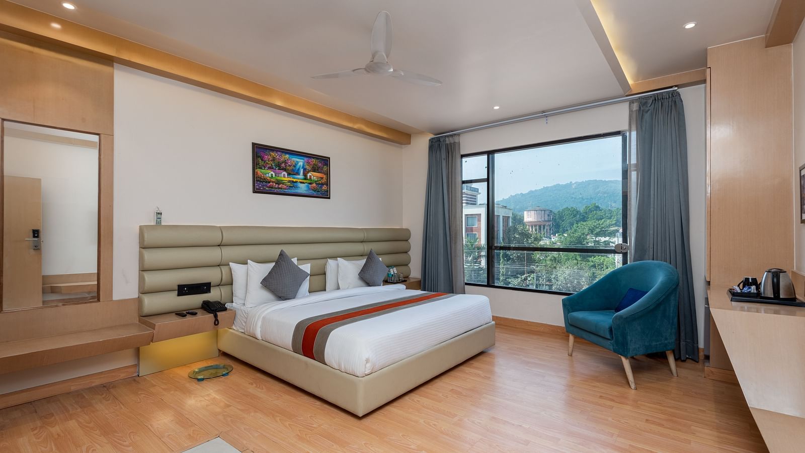 A room with a bed, bed side tables, a chair, and an expansive glass window overlooking the city view - DLS Hotel  Arihant, Haridwar