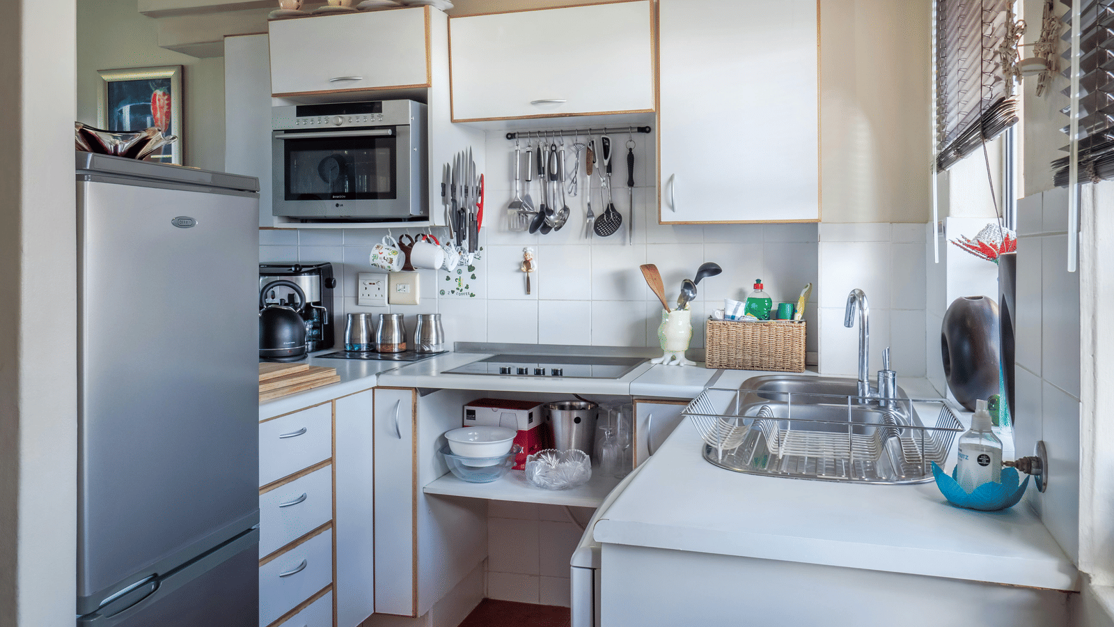 A well-lit, compact kitchen featuring white cabinetry, a stainless steel sink, and various kitchen utensils and appliances.