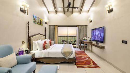a hotel room in candolim featuring a double bed with a fancy headboard, a blue love seat, high ceilings with a fan, and a balcony featuring verdant views - Golden Tulip, Candolim, Goa