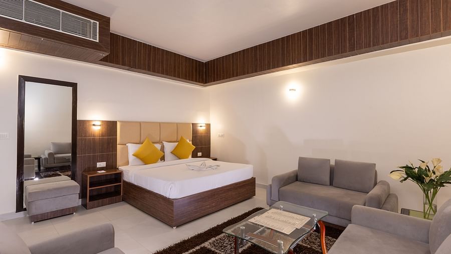A hotel suite or large bedroom featuring a bed, a full-length mirror, and a separate sitting area with a sofa and table - Pride Premier Dehradun