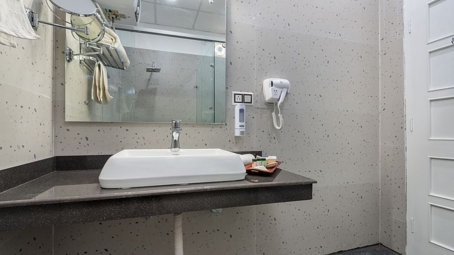 A modern bathroom featuring a rectangular countertop basin, wall-mounted mirror, hairdryer, and a set of toiletries neatly arranged, all set against minimalist grey walls and a dark tiled floor - DLS Nature Trinket Resort, Dalhousie.