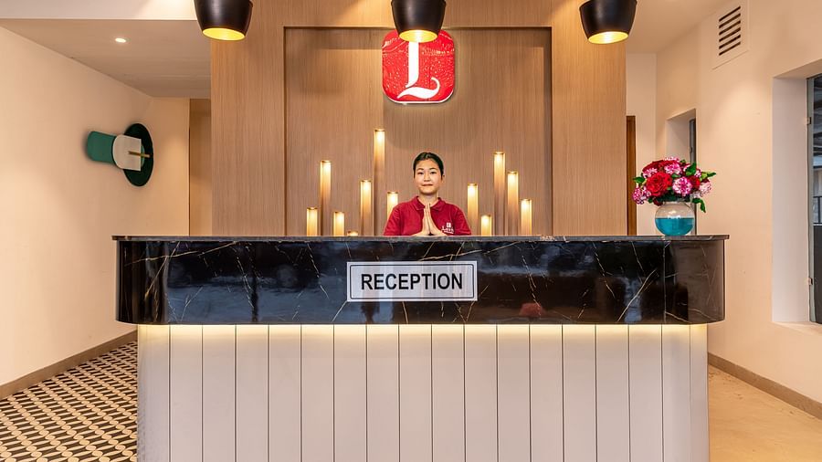 A hotel reception desk with a staff member behind it, featuring hanging pendant lights and a Lords logo. Includes a patterned floor and decorative elements on the desk - Lords Eco Inn Dehradun.