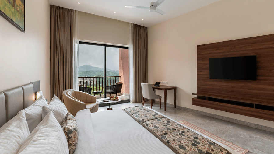 A premium room at Osian Valley Resort & Spa, Kumbhalgarh, featuring a plush bed, large windows with scenic outdoor views, a study desk, and an inviting neutral interior design.