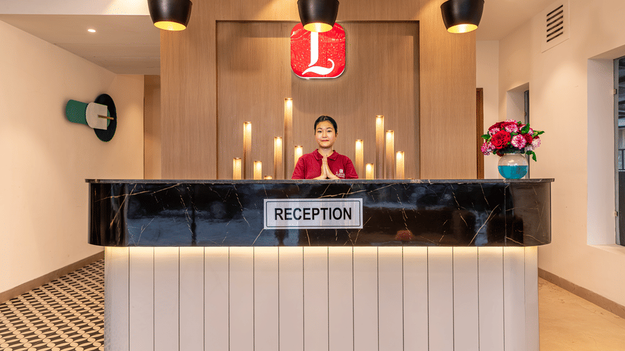 A modern designed reception desk with a staff member welcoming with hand gesture, featuring hanging pendant lights, a patterned floor and decorative elements on the desk at Lords Eco Inn Dehradun.