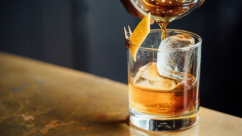 A cocktail being poured onto ice spheres in a glass placed on a bar counter