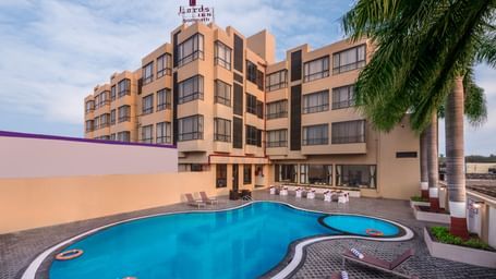 A shot of a building with a swimming pool and some lounge chairs in front.