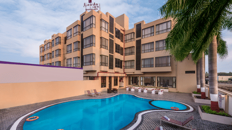 A shot of a building with a swimming pool and some lounge chairs in front.