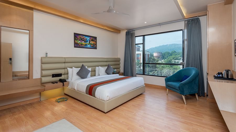 A room with a bed, bed side tables, a chair, and an expansive glass window overlooking the city view - DLS Hotel  Arihant, Haridwar