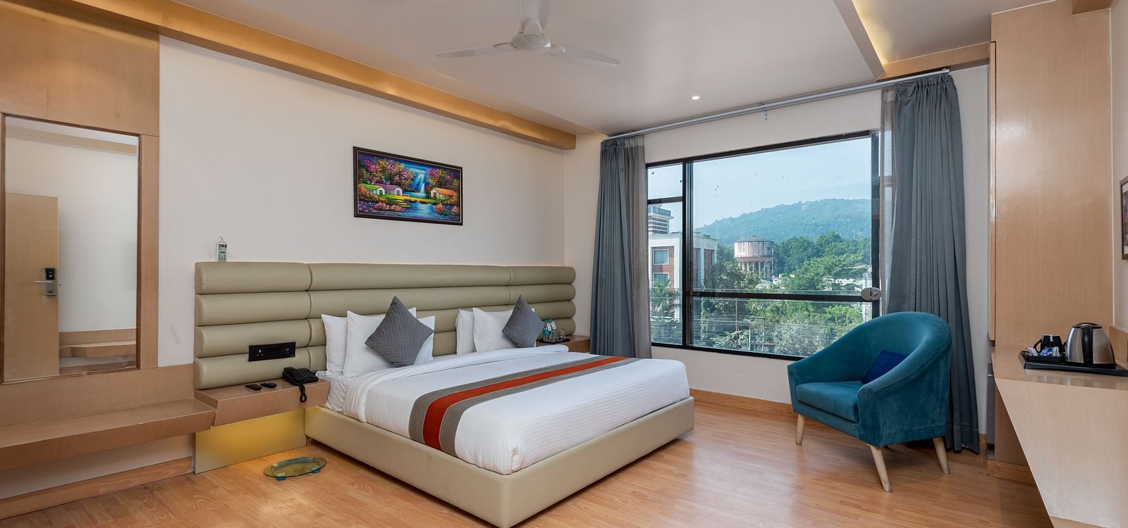 A room with a bed, bed side tables, a chair, and an expansive glass window overlooking the city view - DLS Hotel  Arihant, Haridwar