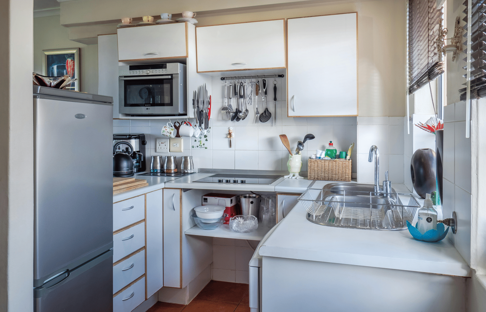 A well-lit, compact kitchen featuring white cabinetry, a stainless steel sink, and various kitchen utensils and appliances.