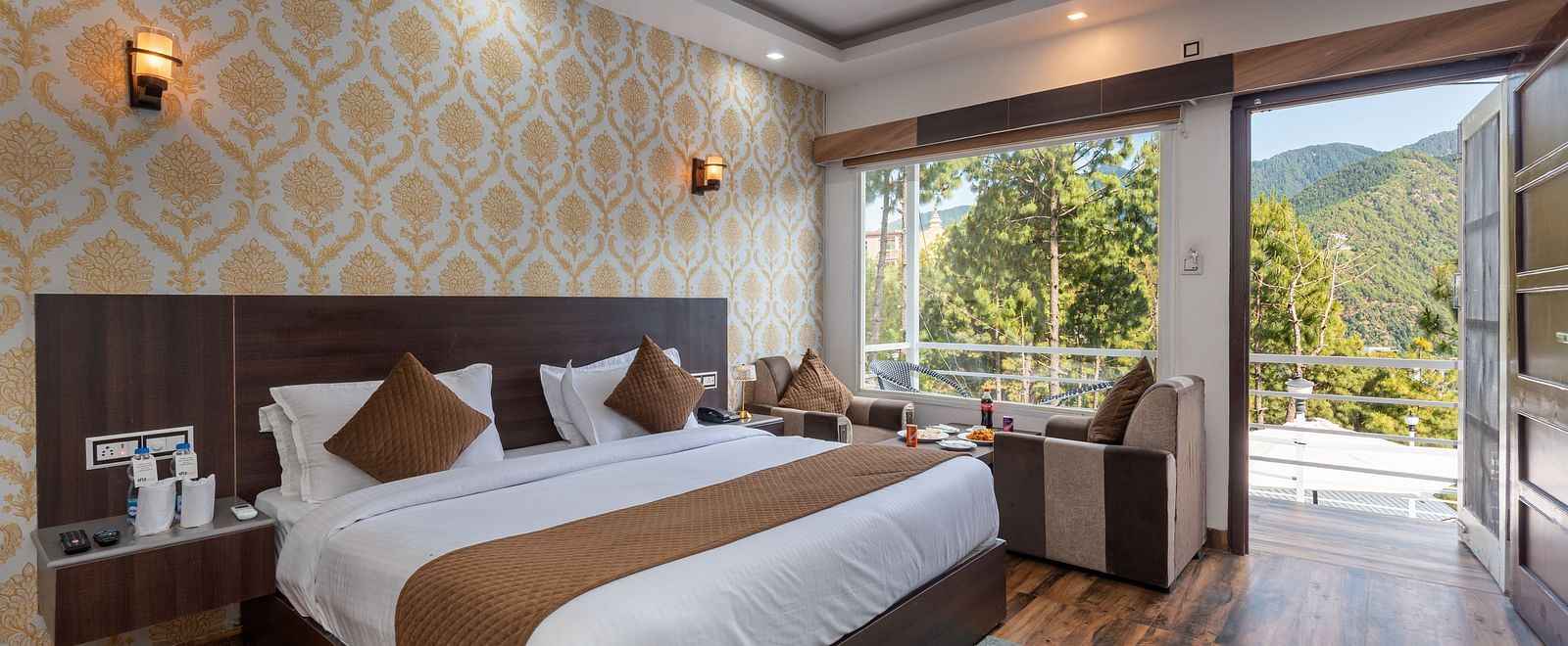  Modern hotel room with a large bed, elegant wallpaper, and wooden flooring; features a seating area, ceiling fan, and a balcony with a scenic mountain view - DLS Nature Trinket Resort, Dalhousie.