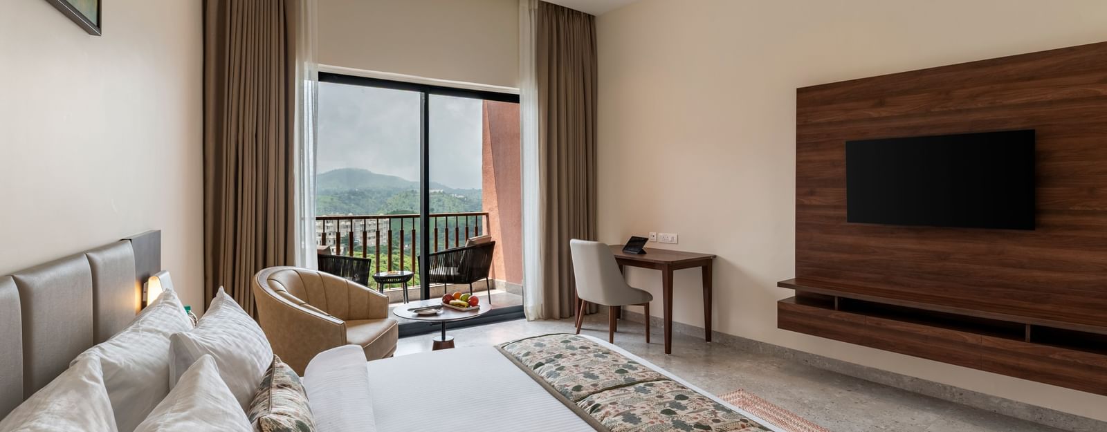 A modern bedroom at Osian Valley Resort & Spa with a large bed, a TV mounted on a wooden wall, and a balcony with a view of a lush green landscape.