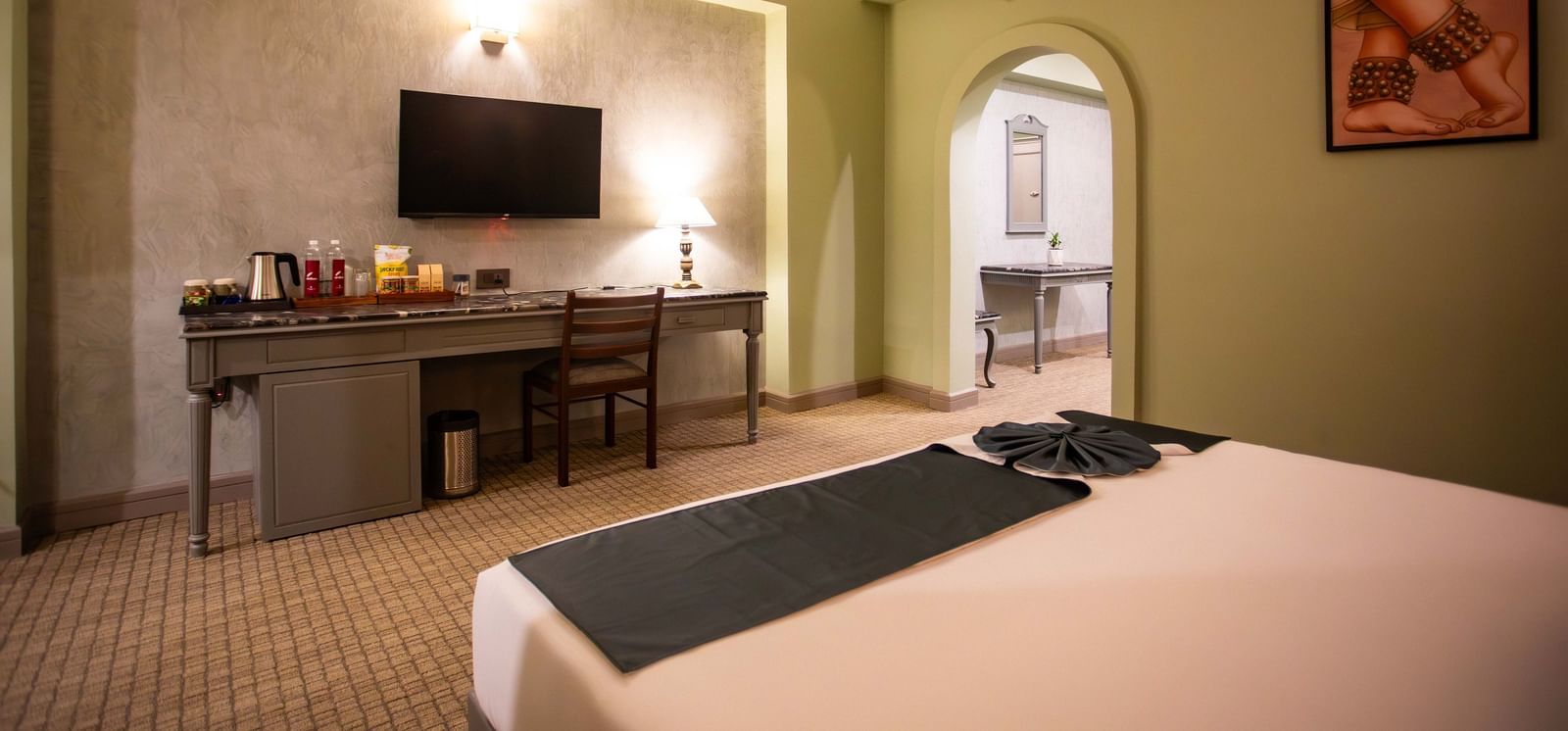  A guest room at Shilton International, Kochi, with a large bed, desk, television, and an archway leading to another area.