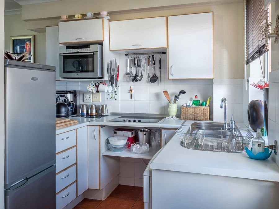 A well-lit, compact kitchen featuring white cabinetry, a stainless steel sink, and various kitchen utensils and appliances.