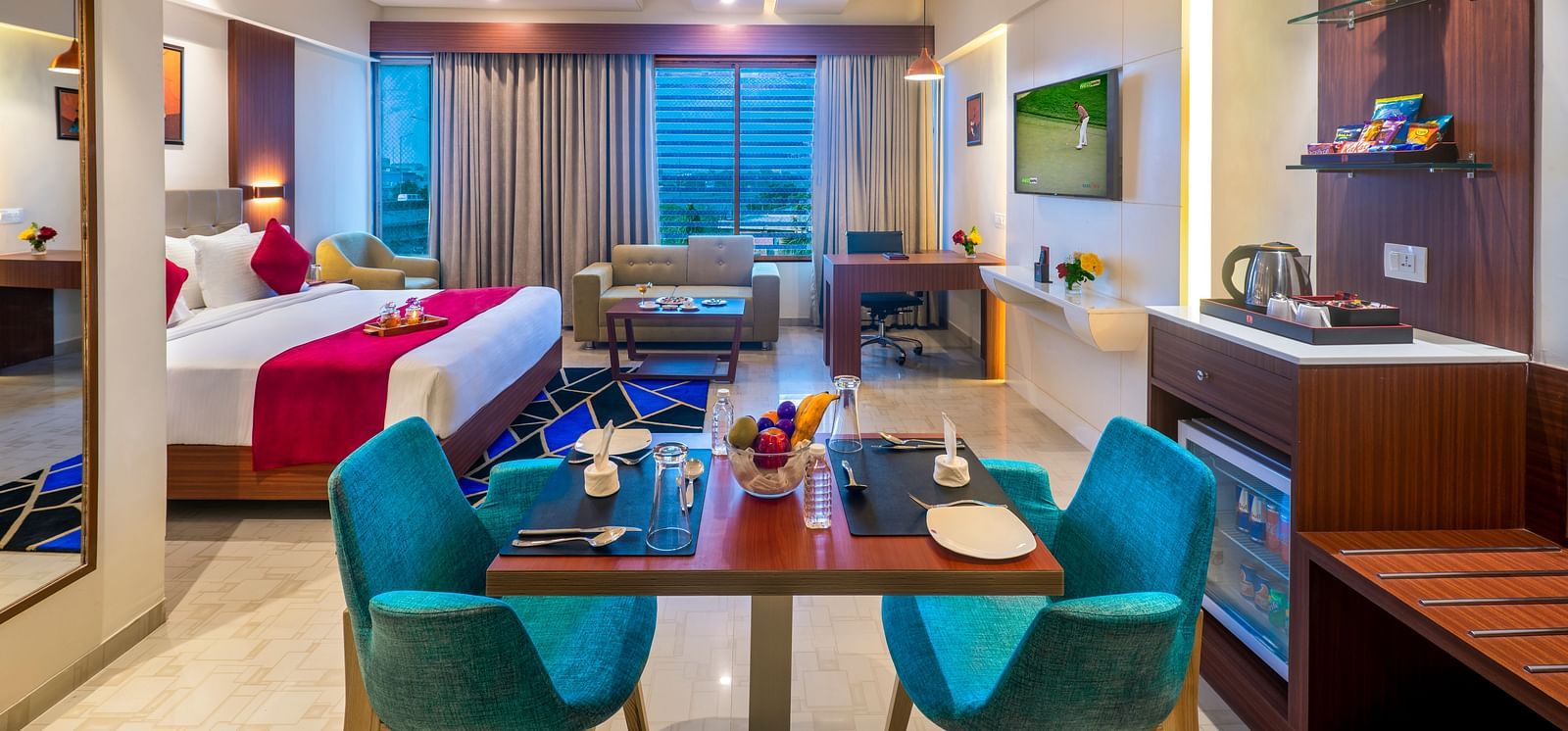 A hotel suite with a bed featuring blue and white bedding, a dining table with blue chairs, and a view of the outside.