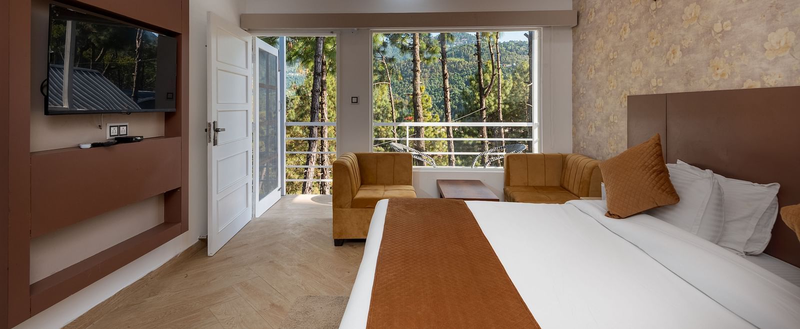  Bright hotel room with a brown and beige theme, featuring a double bed, flat-screen TV, cosy seating area, and large windows overlooking a forested view - DLS Nature Trinket Resort, Dalhousie.