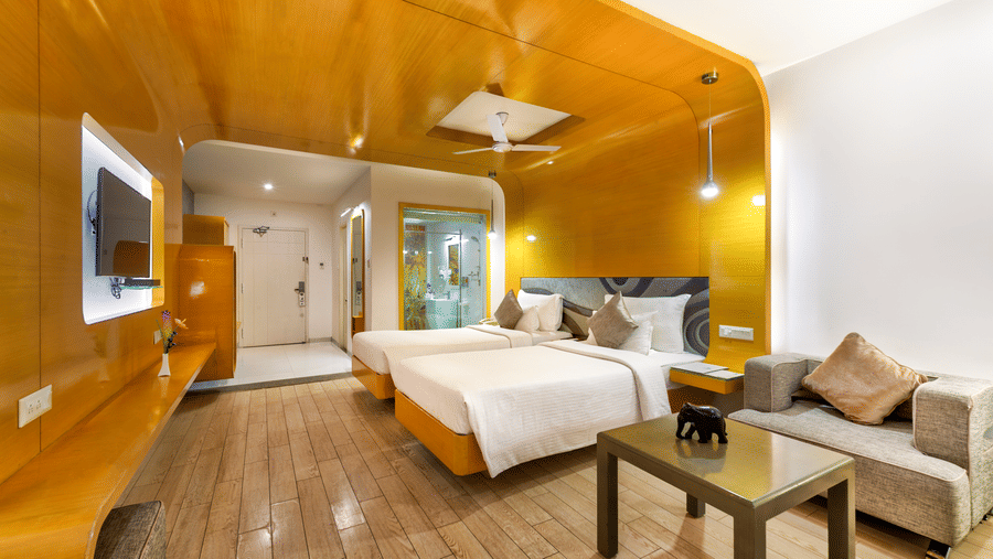 A room with twin beds, golden wall panel and wooden furniture featuring a seating space for a comfortable stay - Mahabalipuram's Grande Bay Resort & Spa.