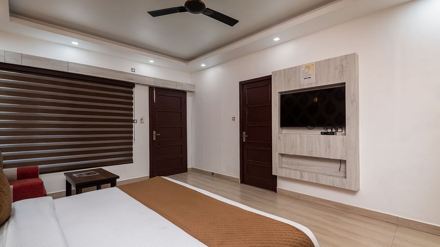  A modern hotel room featuring a neatly made double bed with brown and white linens, a wall-mounted flat-screen TV, wooden flooring, a ceiling fan, and a window with brown blinds - DLS Nature Trinket Resort, Dalhousie.
